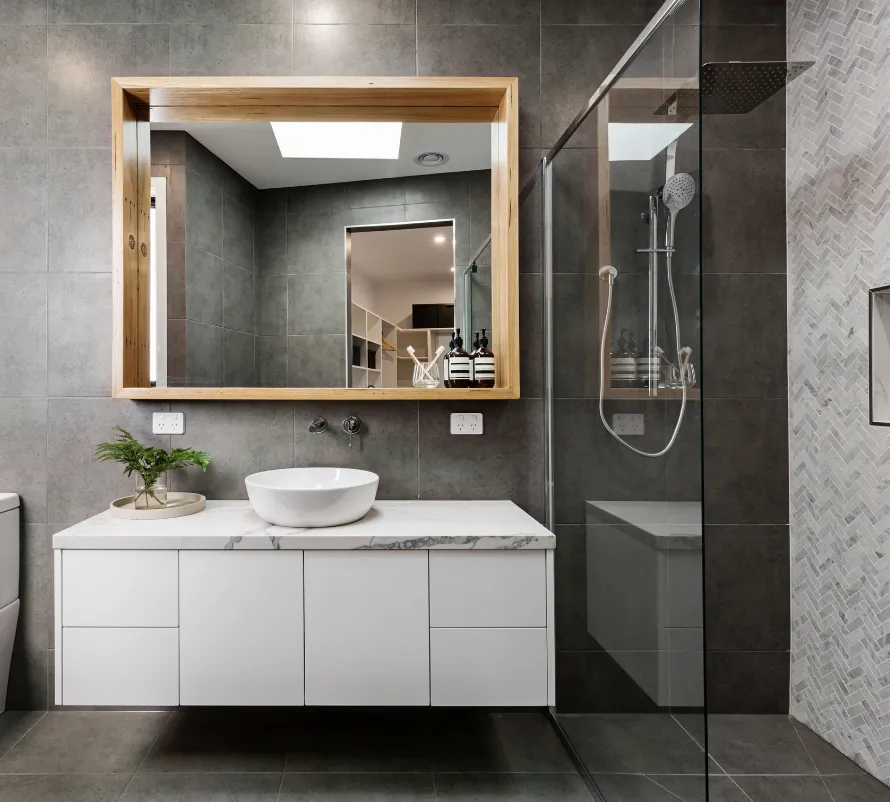 bathroom-1 Modern bathroom with gray tiles, a glass shower, white vanity, vessel sink, and large mirror with wood frame.