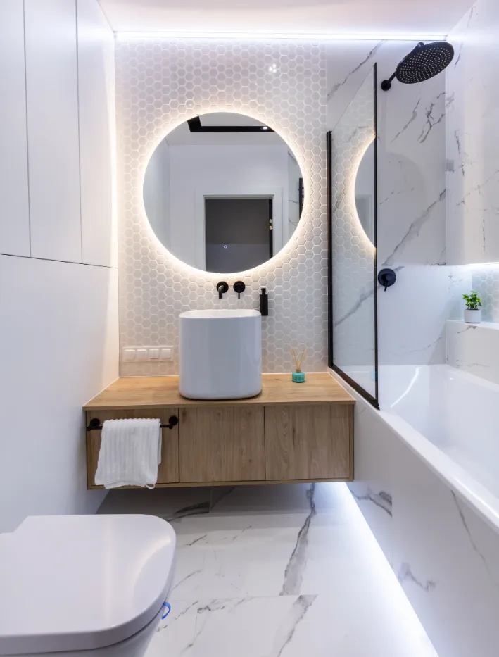 bathroom-3 Modern bathroom with round backlit mirror, vessel sink, wooden vanity, and marble walls and floor.