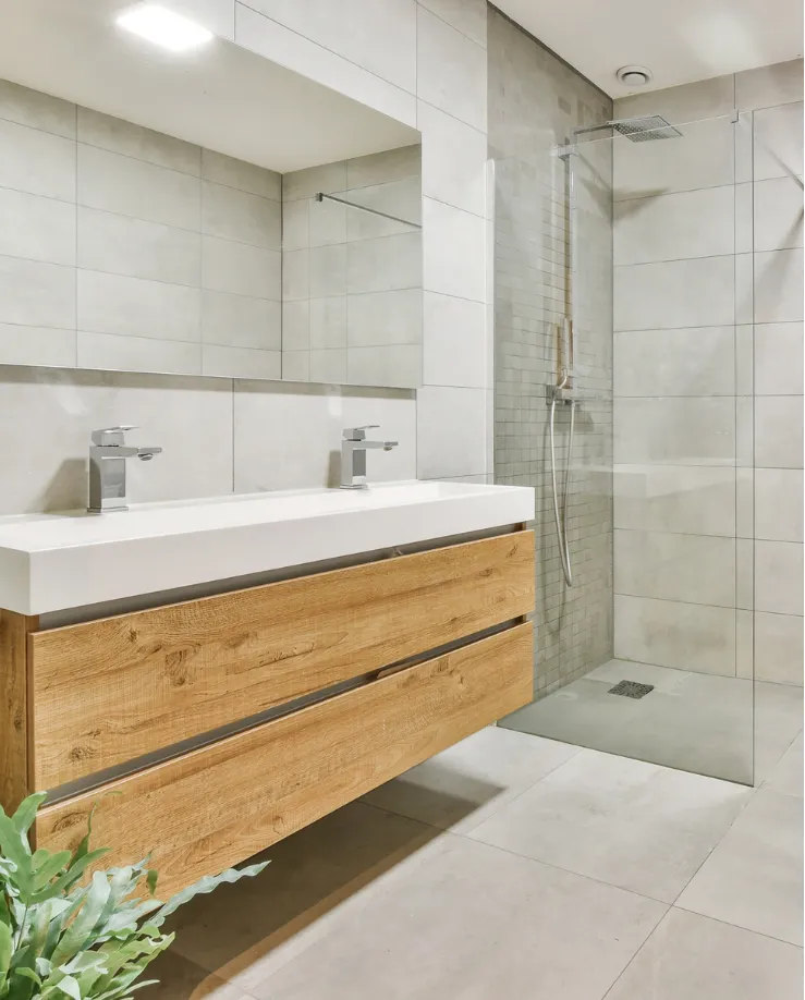 bathroom-4 Modern bathroom with a wooden vanity, double sink, glass shower, and light gray tile walls and floor.