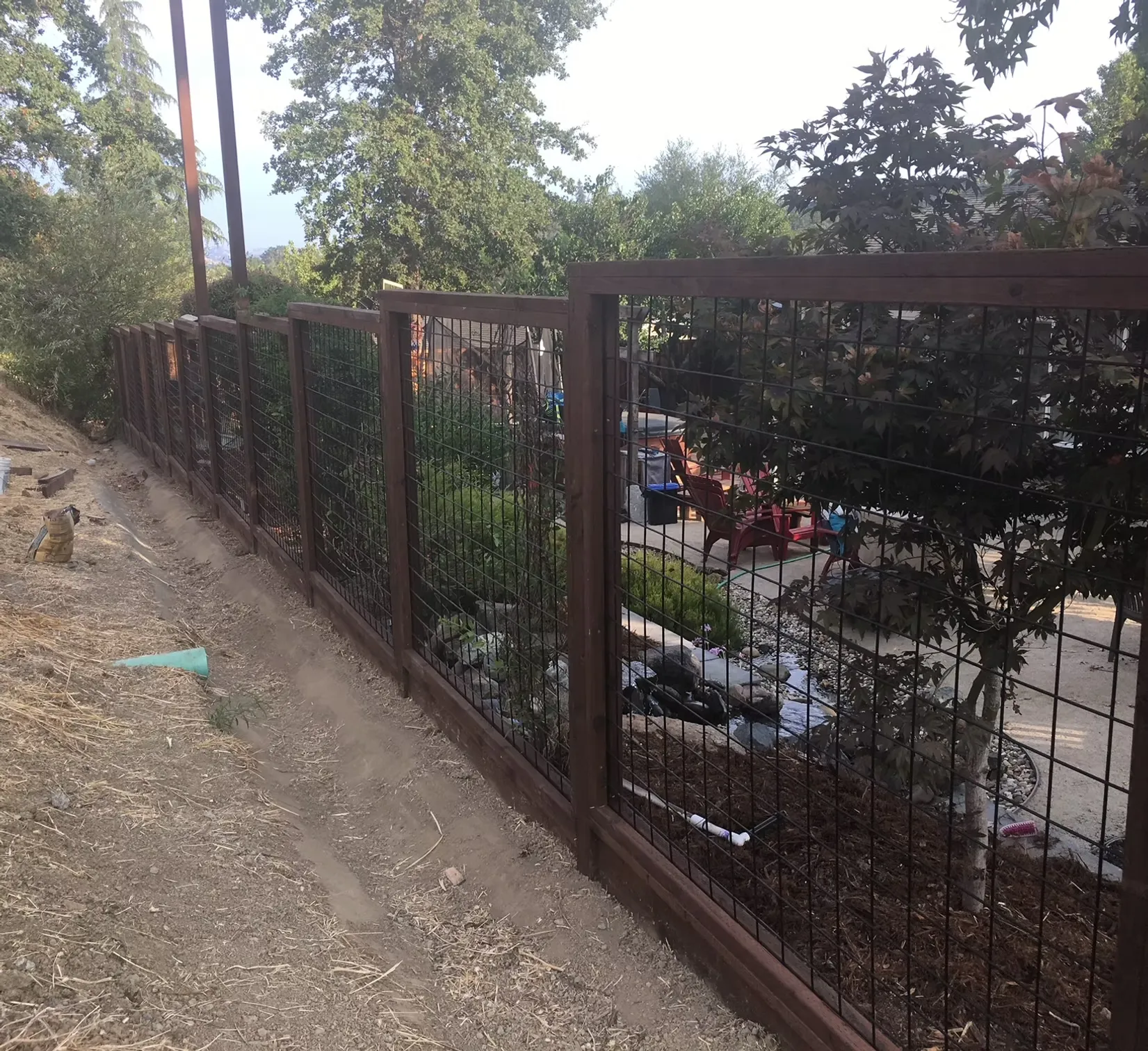 welcome-1 A wire and wood fence runs along a sloped dirt yard beside bushes and a driveway with parked cars.