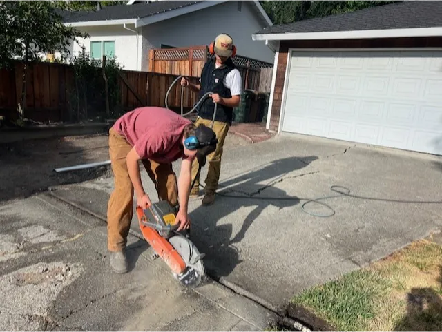 construction-7 Two workers use a concrete saw to cut a driveway near a garage on a sunny day.