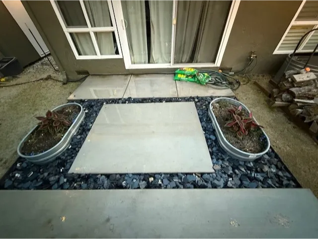 construction-9 Square patio with gray tiles, dark rocks, and two metal planters with small plants near glass doors.