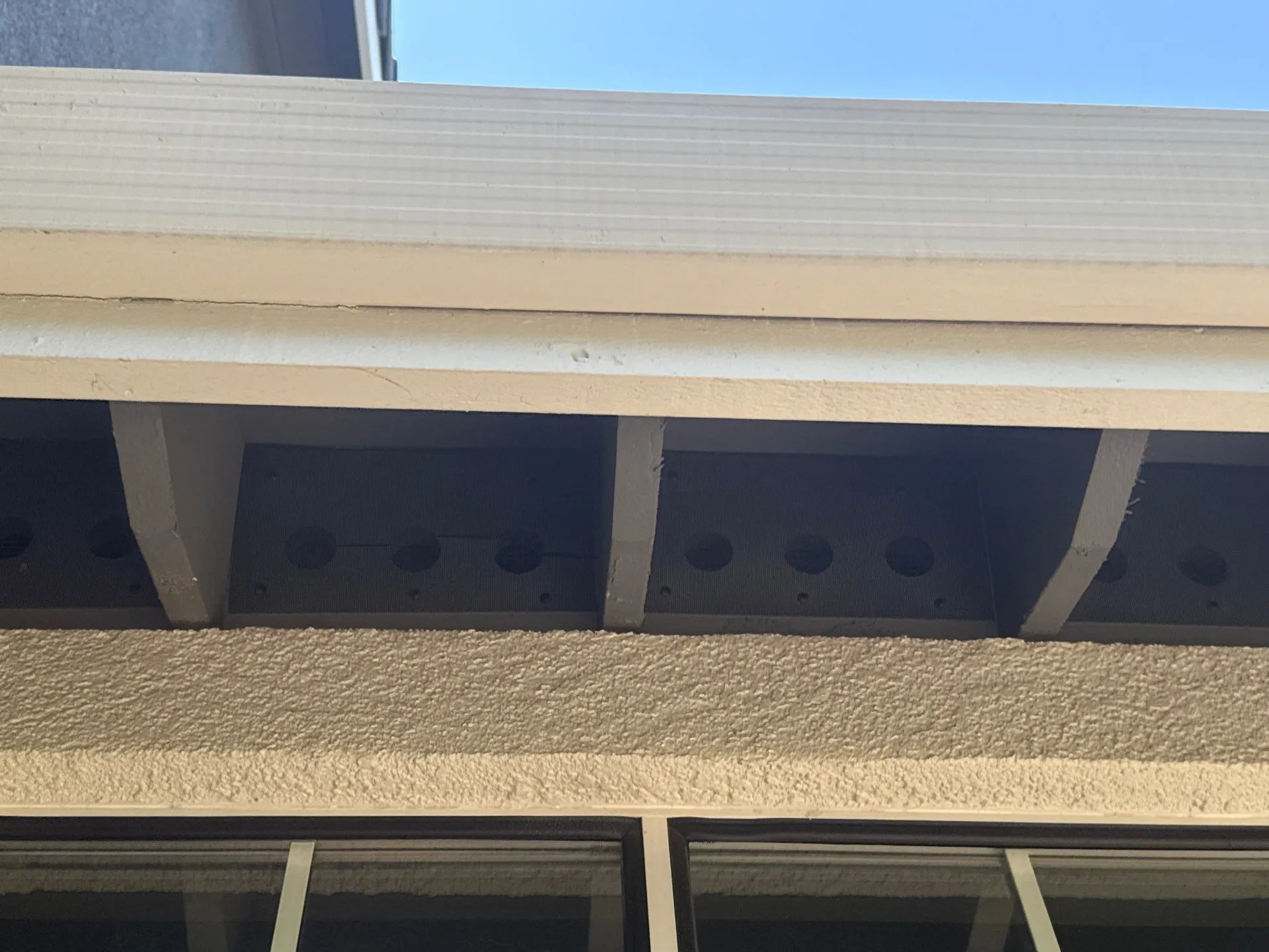 hardening-experts-2 Close-up of a beige roof eave with vented soffit panels and a clear blue sky above.