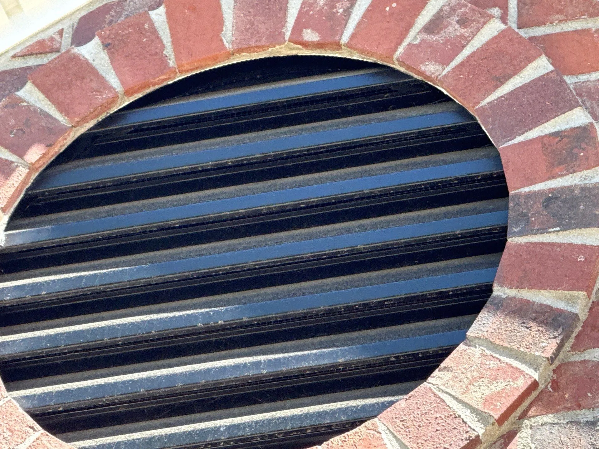 hardening-experts-3 Round brick vent with black metal slats viewed at an angle, showing brickwork and louvered grill.